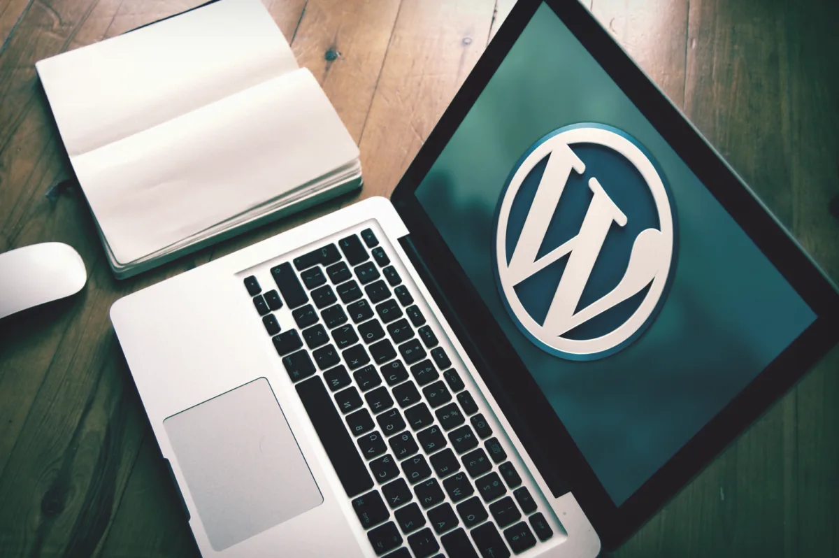 Laptop z logo WordPressa na ekranie i otwarty notes na drewnianym biurku